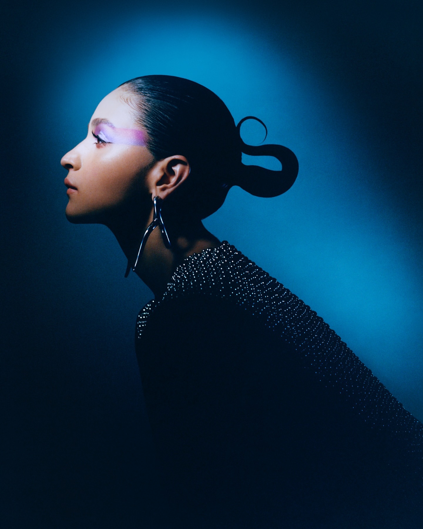 Profile of a woman with creative hair, wearing an embellished top and large earrings against a blue gradient background in a beauty shot.