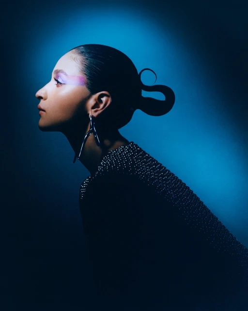 Profile of a woman with creative hair, wearing an embellished top and large earrings against a blue gradient background in a beauty shot.