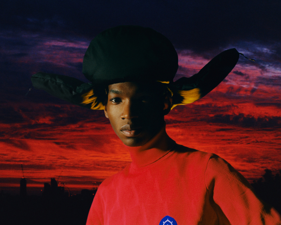 Portrait of adult male in red turtleneck and oversized hat against sunset skyline backdrop, medium shot, portrait orientation.