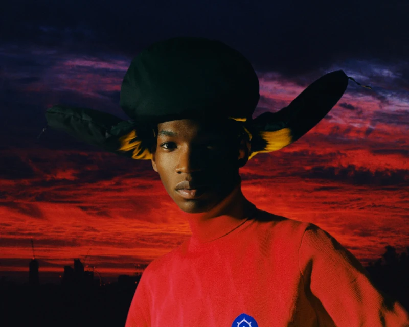 Portrait of adult male in red turtleneck and oversized hat against sunset skyline backdrop, medium shot, portrait orientation.
