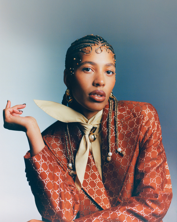 Portrait of a woman with braids wearing an orange patterned blazer and a cream colored tie. She's posing in front of a gradient blue background.
