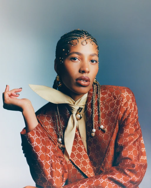 Portrait of a woman with braids wearing an orange patterned blazer and a cream colored tie. She's posing in front of a gradient blue background.