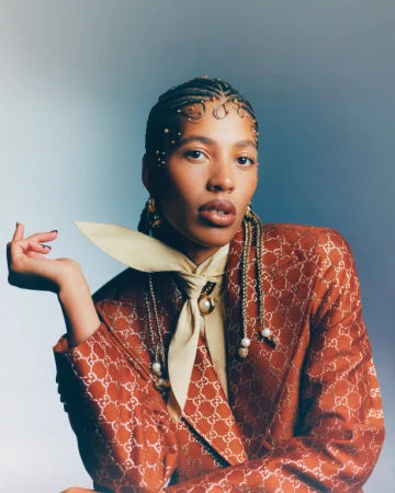 Portrait of a woman with braids wearing an orange patterned blazer and a cream colored tie. She's posing in front of a gradient blue background.