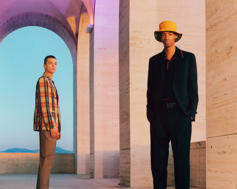 Two men in fashionable clothing stand within an arched colonnade with light blue sky, showcasing modern style and architectural setting.
