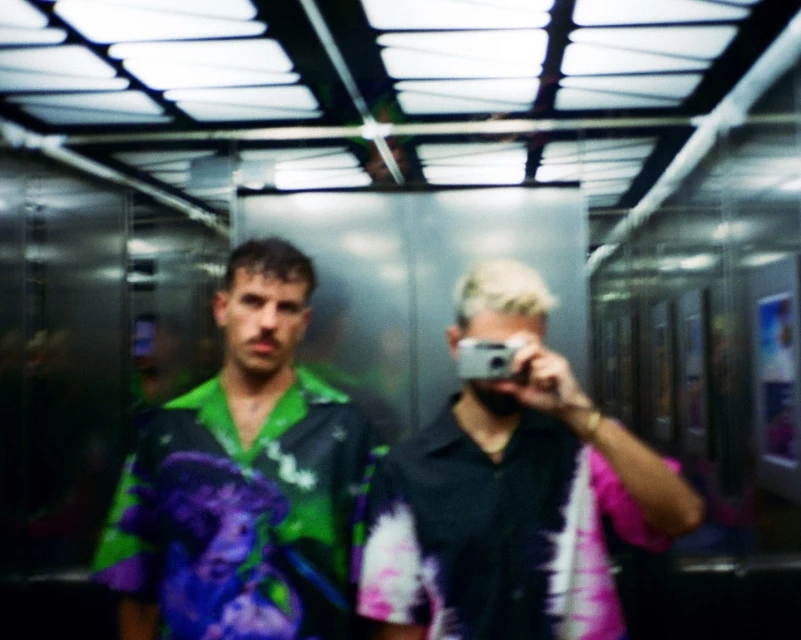 Two adult men in an elevator, one taking a photo with a digital camera. The image is captured at eye-level with indoor lighting.