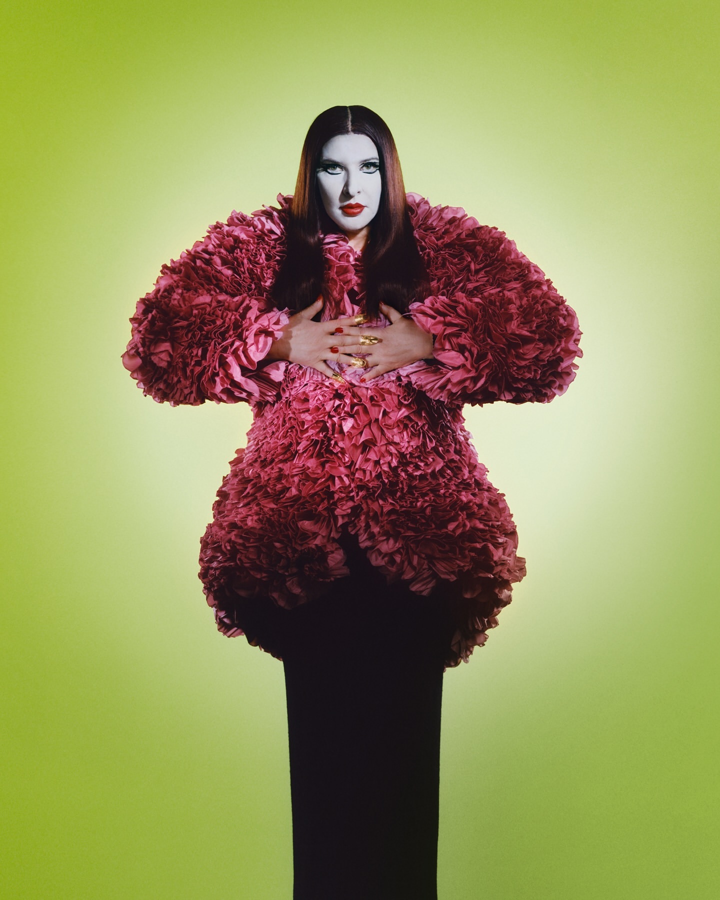 Adult female fashion model in a large, ruffled pink coat over a black gown, posing in front of a green gradient background.