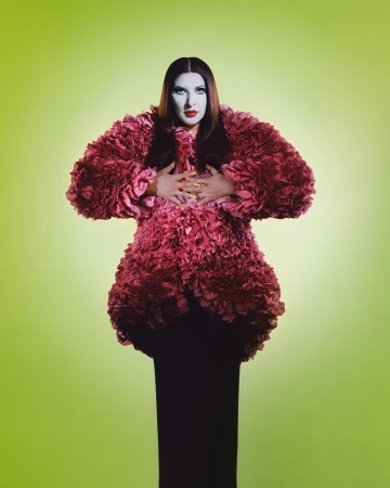 Adult female fashion model in a large, ruffled pink coat over a black gown, posing in front of a green gradient background.