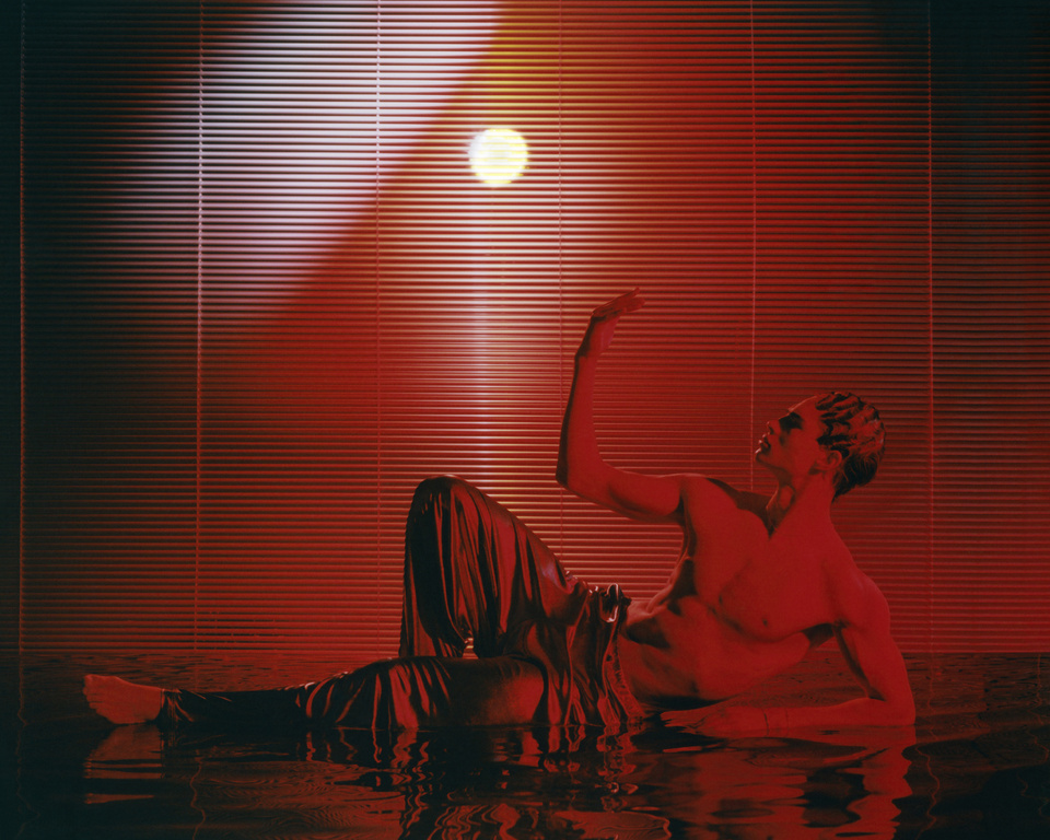 Artistic studio shot of a fair-skinned adult male reclining with his arm raised, under red light and window blinds.