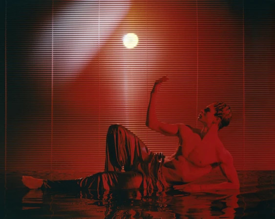 Artistic studio shot of a fair-skinned adult male reclining with his arm raised, under red light and window blinds.