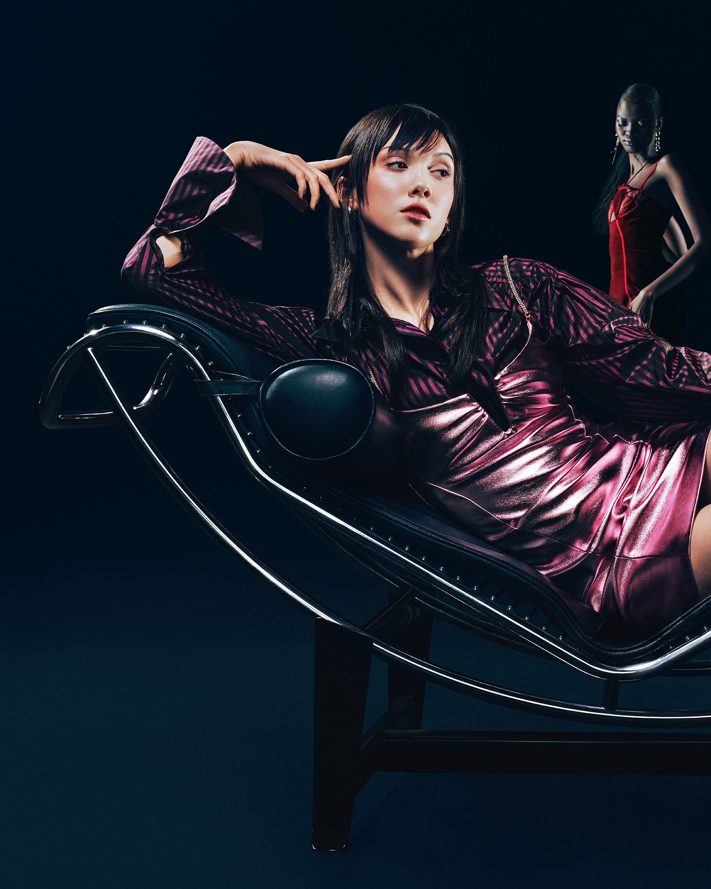 Elegant woman in a magenta dress poses on a chaise lounge with a blurred woman in red standing in the dark background.