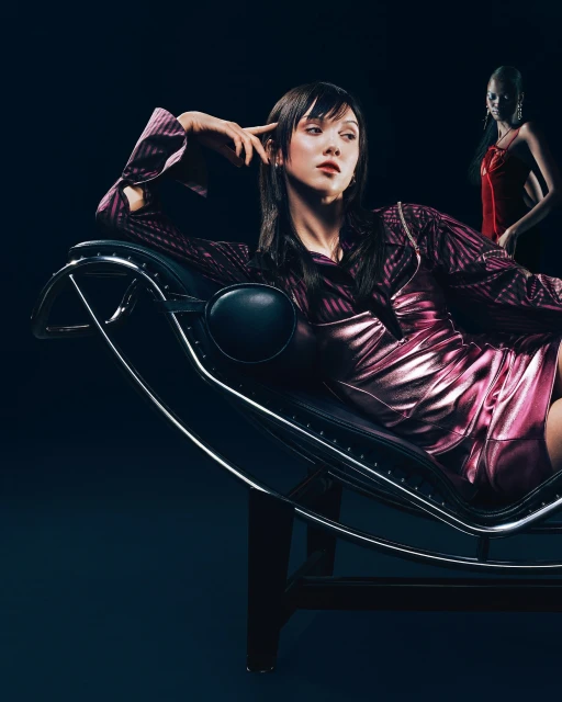 Elegant woman in a magenta dress poses on a chaise lounge with a blurred woman in red standing in the dark background.