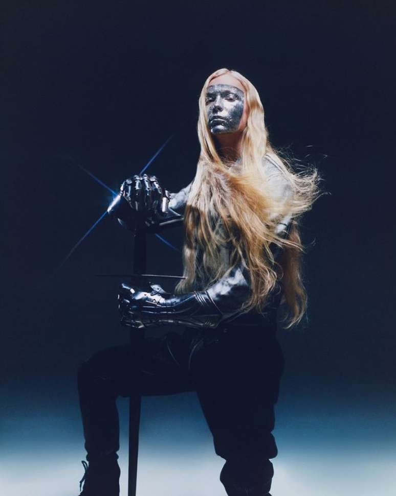 A person with long, light-colored hair and metallic silver makeup poses with a sword in a dramatic, dark studio shot. 