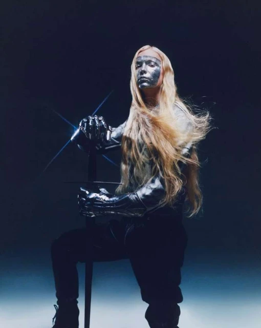 A person with long, light-colored hair and metallic silver makeup poses with a sword in a dramatic, dark studio shot. 