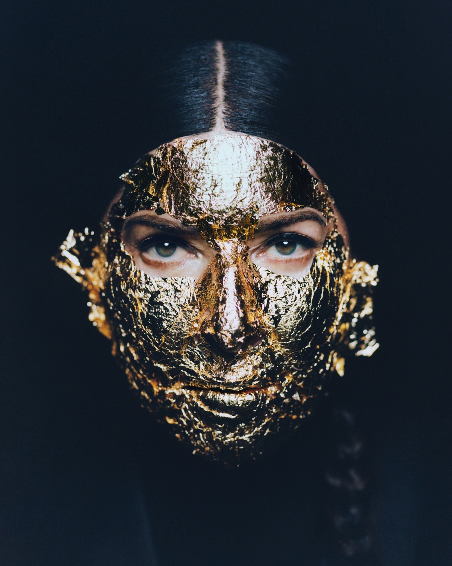 Close-up of a female portrait with a golden leaf mask covering her face, eyes visible against a dark background. Beauty concept.