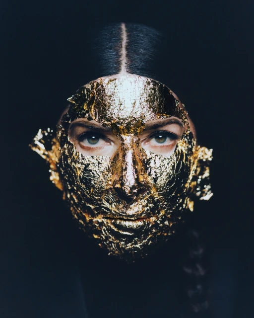 Close-up of a female portrait with a golden leaf mask covering her face, eyes visible against a dark background. Beauty concept.