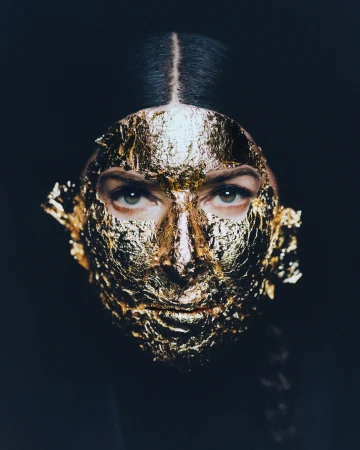 Close-up of a female portrait with a golden leaf mask covering her face, eyes visible against a dark background. Beauty concept.