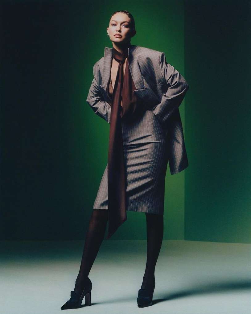 Adult woman in a pinstripe suit stands in front of a green backdrop wearing stockings and heels while posing for a fashion shot.