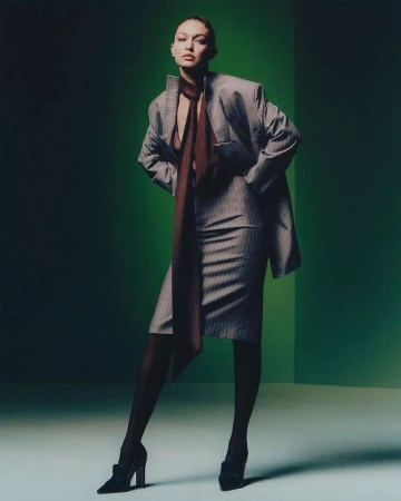 Adult woman in a pinstripe suit stands in front of a green backdrop wearing stockings and heels while posing for a fashion shot.