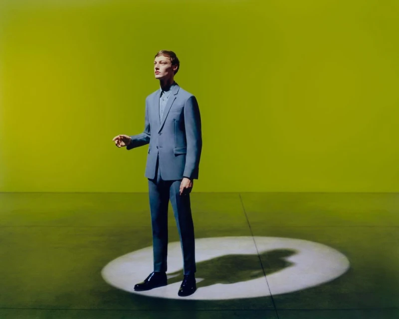 An adult male wearing a blue suit standing in the center of a spotlight against a yellow-green backdrop looking off into the distance.