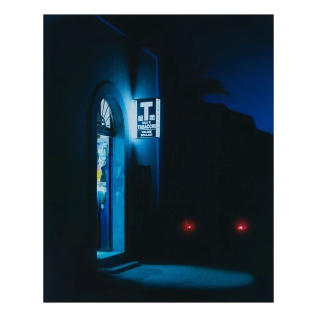 Night scene of an Italian tobacco shop exterior with illuminated signage, and a glimpse of the interior through the open arched doorway.