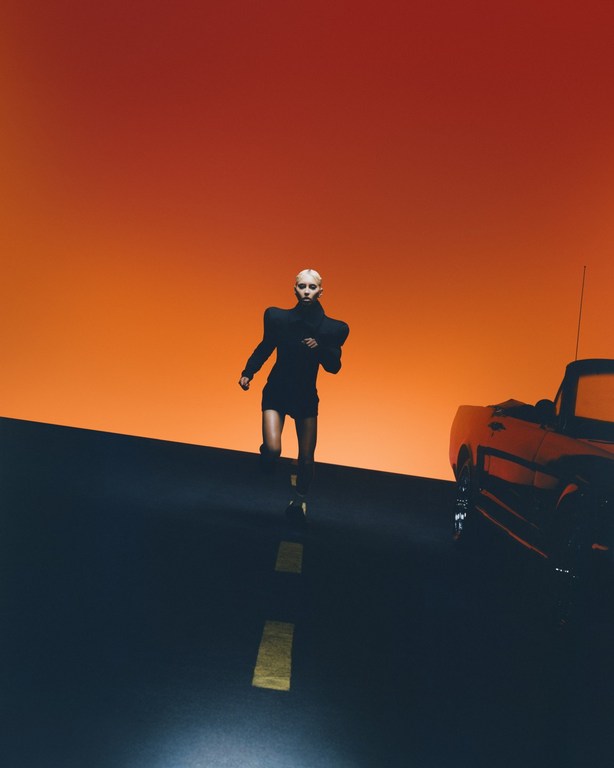 Model in black structured shoulders outfit running on road beside car with orange sunset sky