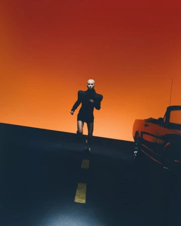 Model in black structured shoulders outfit running on road beside car with orange sunset sky