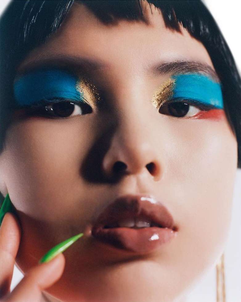 Close-up studio portrait of an Asian female model wearing bold teal and gold eyeshadow with glossy taupe lips and green fingernails.