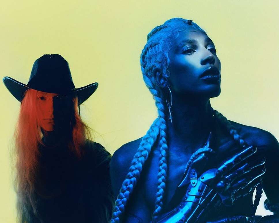 Two figures stand against a yellow backdrop. One wears a cowboy hat; the other with light blue braided hair and a metallic hand piece.