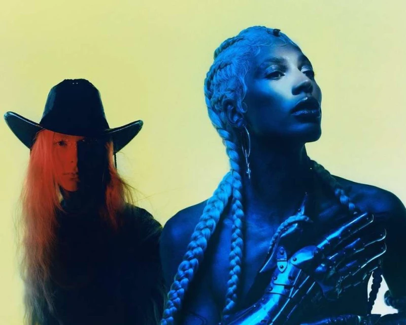 Two figures stand against a yellow backdrop. One wears a cowboy hat; the other with light blue braided hair and a metallic hand piece.