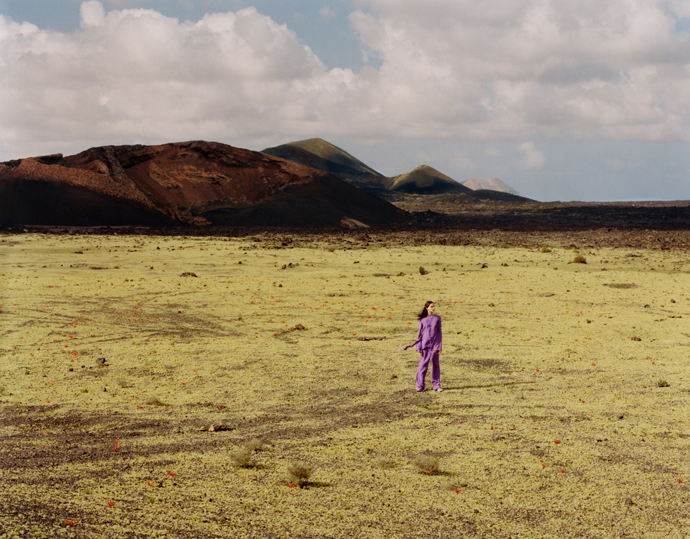 A woman in a purple outfit stands in a barren landscape with mountains under a cloudy sky, creating a striking contrast and scenic view.