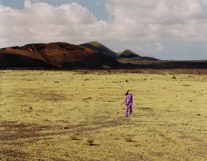 A woman in a purple outfit stands in a barren landscape with mountains under a cloudy sky, creating a striking contrast and scenic view.