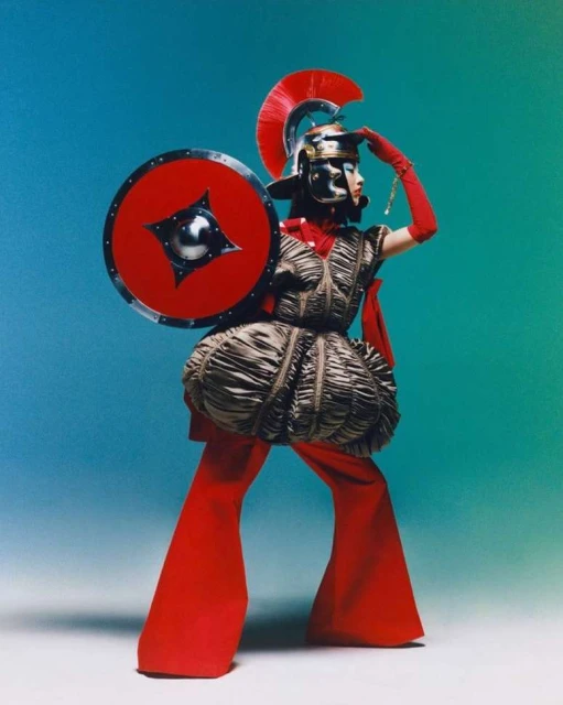 Person in red gladiator-inspired outfit with helmet, shield, and flared pants poses against a vibrant turquoise and green background.