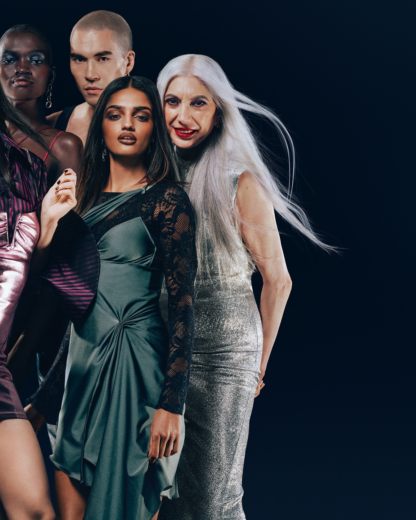 A diverse group of models pose in an indoor studio setting showcasing evening wear in a fashion editorial shot, with moody lighting.