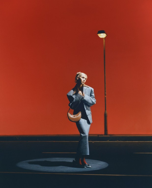 Fashion shot of a fair-skinned woman in a blue suit with a handbag, posing near a street lamp against a bold red background.