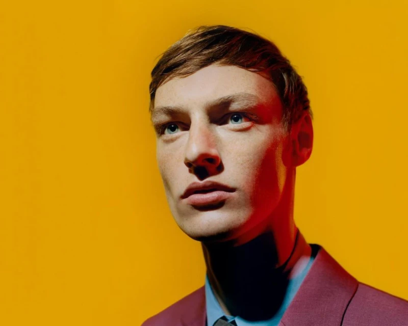 Portrait of a young adult male in a magenta suit with light-brown hair, blue eyes, and fair skin, set against a vibrant yellow background.
