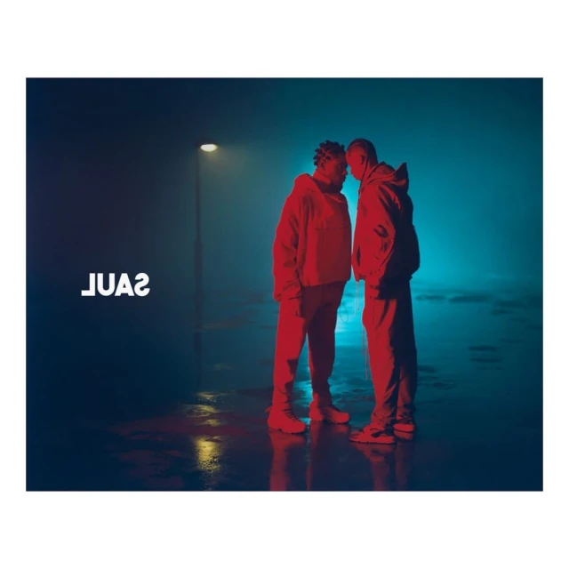 Two people in hooded sweatshirts face each other in a dimly lit, wet urban environment with a street lamp in the background.