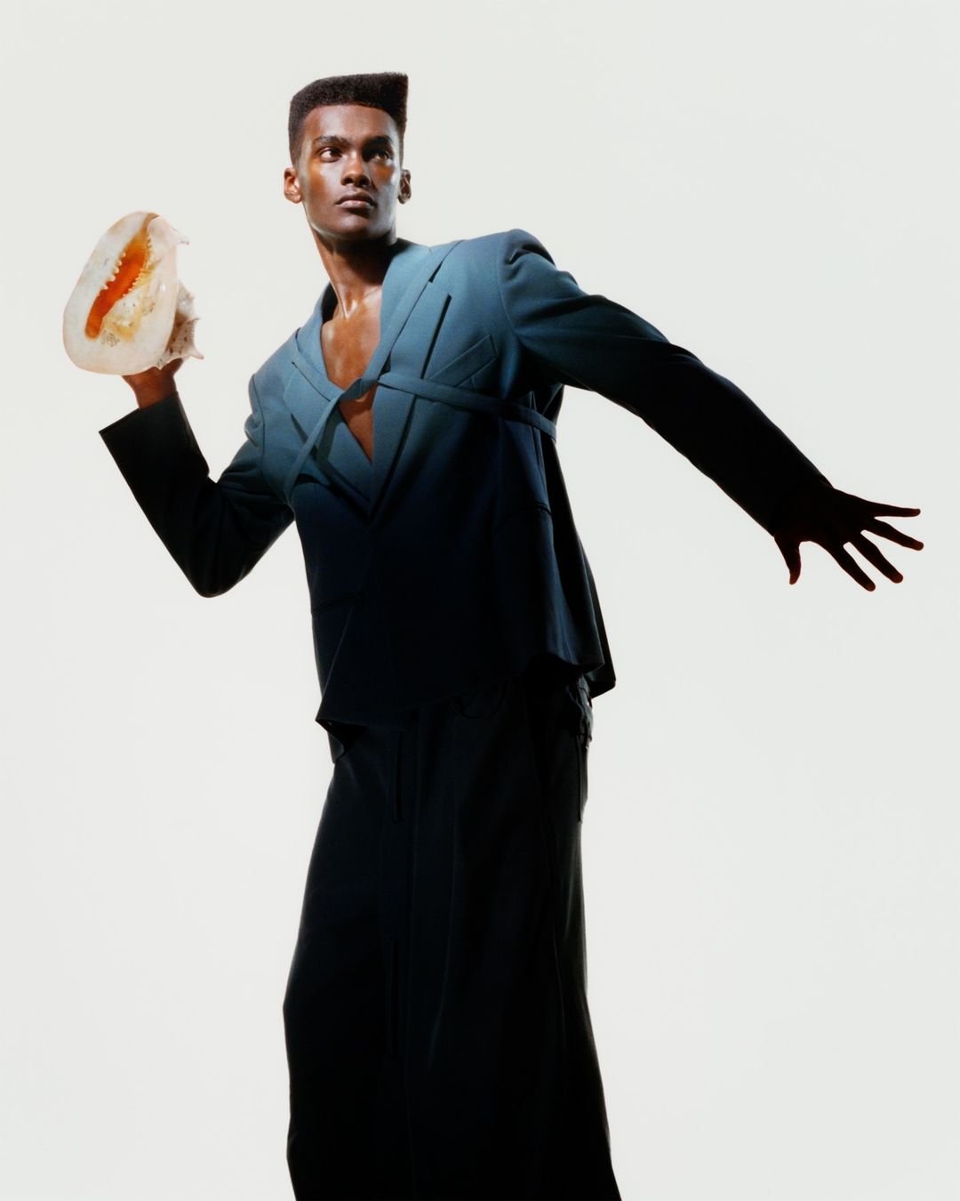 Dark-skinned male fashion model in stylish teal suit and wide-leg trousers poses with a jellyfish against a white background.