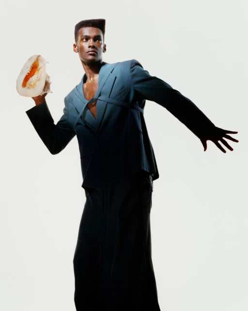 Dark-skinned male fashion model in stylish teal suit and wide-leg trousers poses with a jellyfish against a white background.