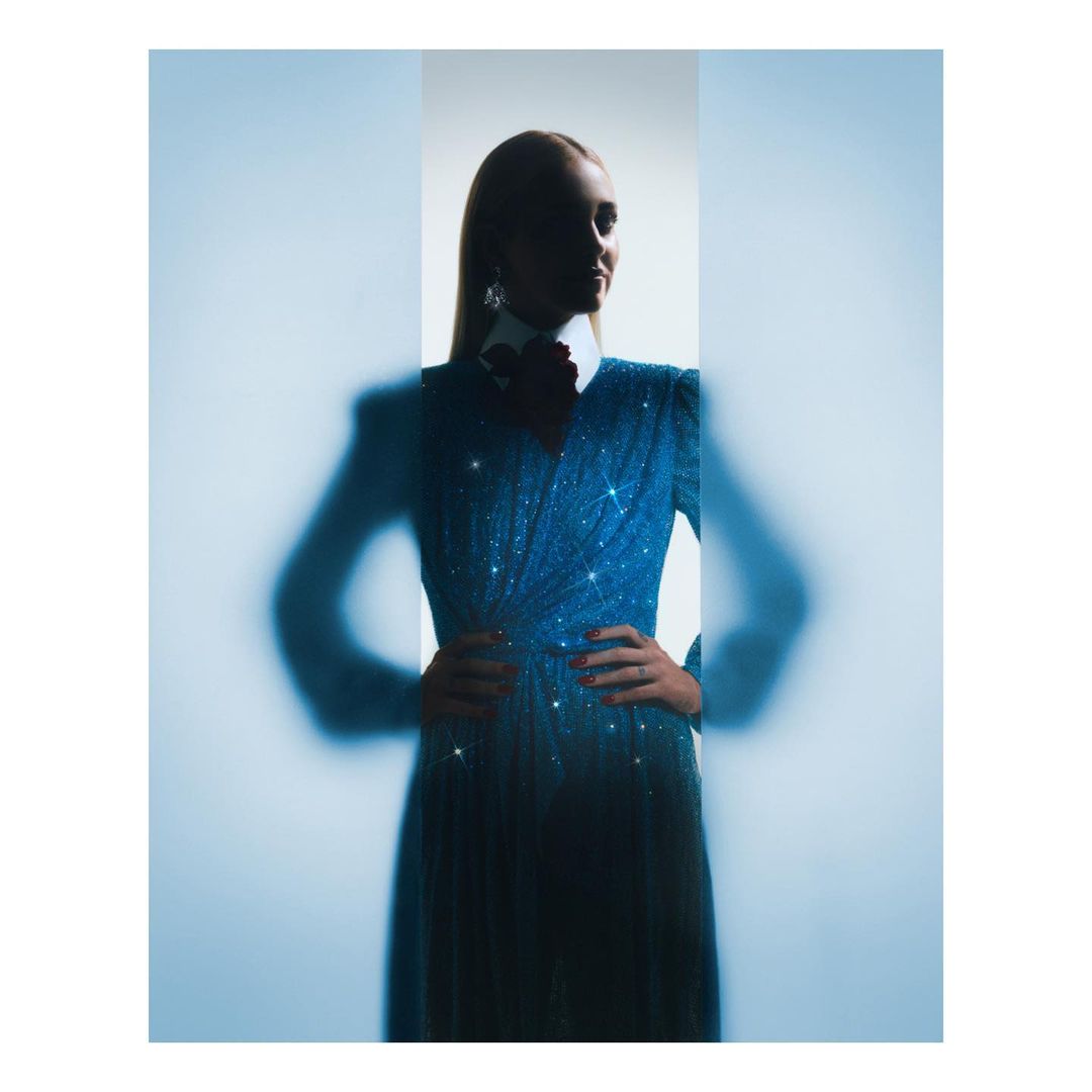 Elegant adult female in a glittering blue gown with an accentuated waistline poses indoors with hands on her hips, soft focus.