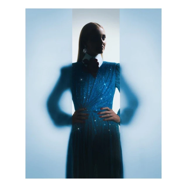 Elegant adult female in a glittering blue gown with an accentuated waistline poses indoors with hands on her hips, soft focus.
