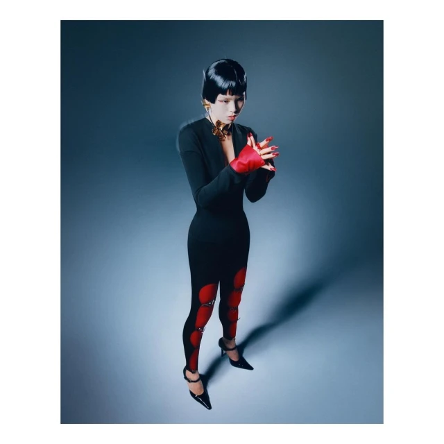A woman with a black bob hairstyle poses in a black dress with red accents and black heels in a studio with a gray gradient background.