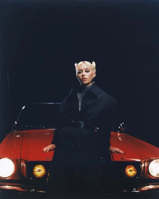 Woman with platinum blonde hair poses on a red vintage car in a studio with dark lighting, wearing a black coat with a belt.