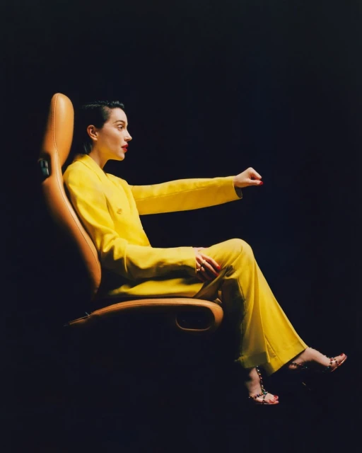 Profile shot of a woman in a yellow suit sitting in a tan-colored chair against a dark background, looking off to the right.