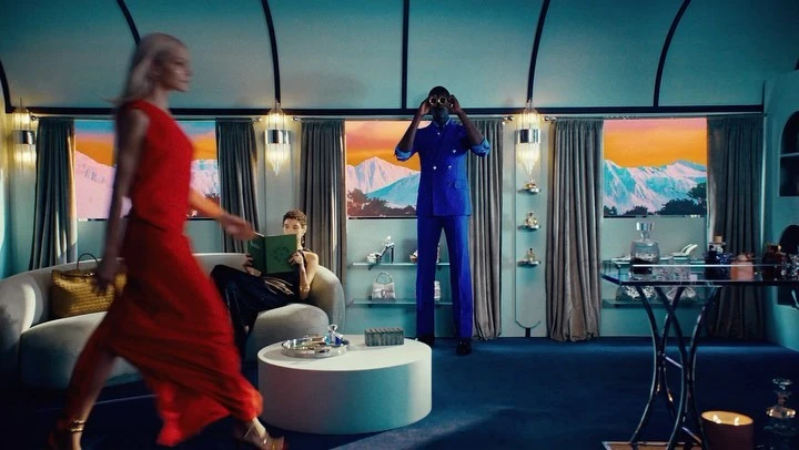 A woman in a red dress walks through a luxurious train car while other passengers relax amid upscale decor and mountain views.
