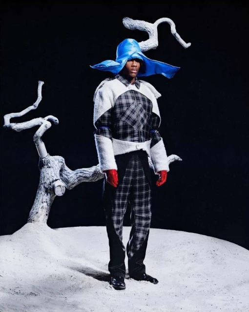 A man stands on a white surface wearing a black and white checkered suit, red gloves, and a large blue hat, next to tree branches.