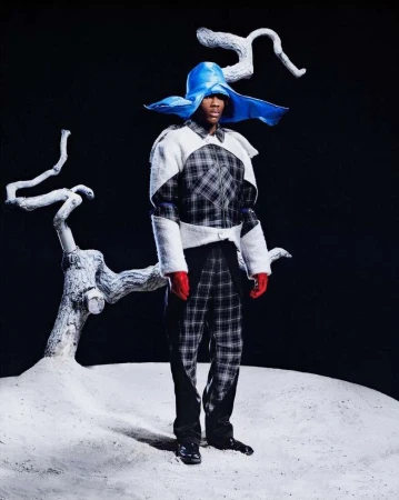 A man stands on a white surface wearing a black and white checkered suit, red gloves, and a large blue hat, next to tree branches.