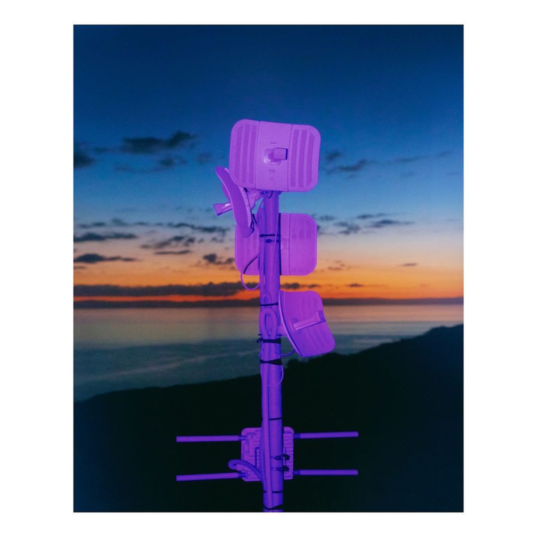 Telecommunications tower stands against a vibrant sunset sky. A mountain range and sea are visible in the distance.
