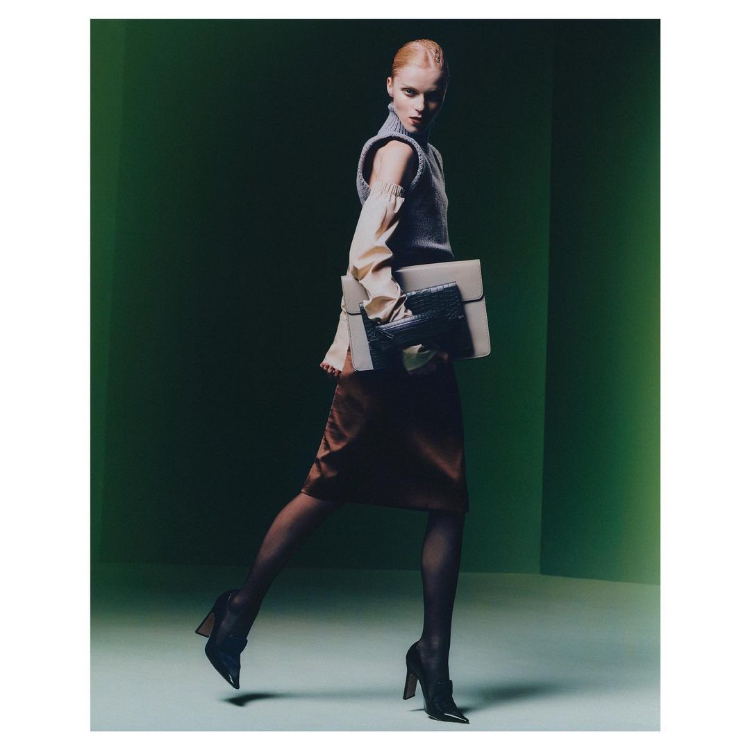 Fashionable woman walking in a studio with handbag and laptop, wearing a sweater vest, long-sleeved shirt, skirt, and heels.