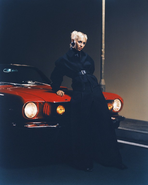 Stylish woman poses against a red vintage car at night, wearing a black coat and chunky earrings under a streetlight, portrait view.