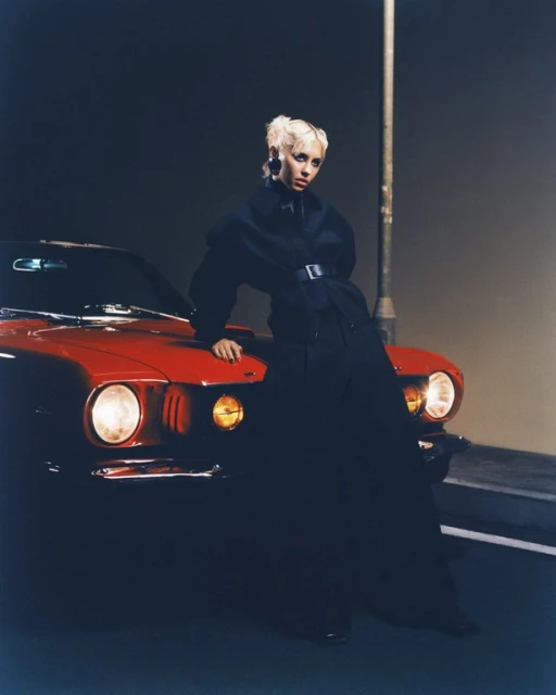 Stylish woman poses against a red vintage car at night, wearing a black coat and chunky earrings under a streetlight, portrait view.